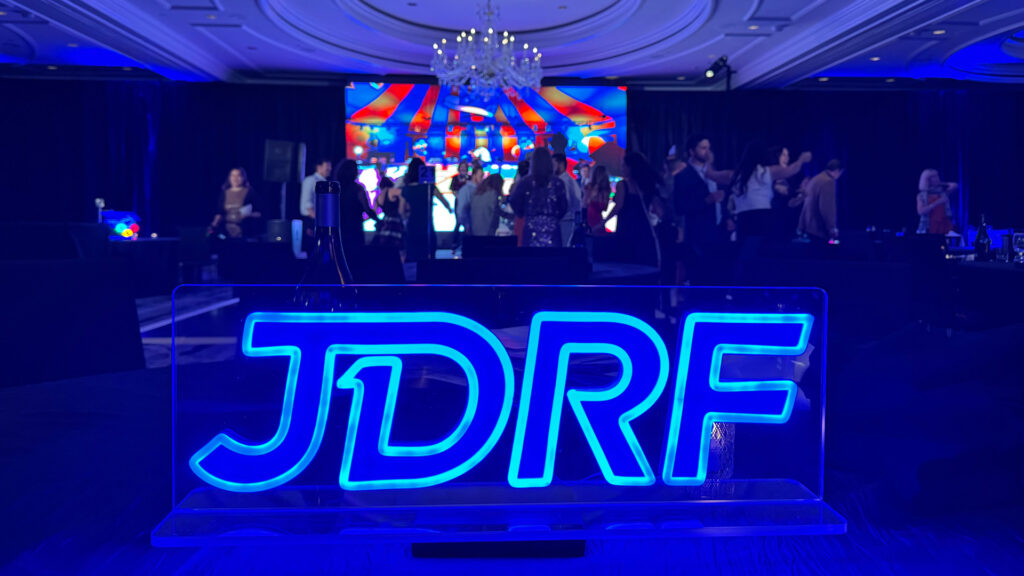 Neon blue JDRF sign in the foreground, with people on a ballroom dance floor in the background.