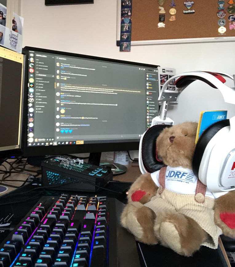 Rufus, the Bear with Diabetes, wearing oversized headphones and sitting in front of a monitor with the JDRF Game2Give Discord on the screen..